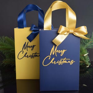 May include: Two gift bags with gold handles and bows. One bag is yellow with a navy blue "Merry Christmas" inscription. The other bag is navy blue with a gold "Merry Christmas" inscription.
