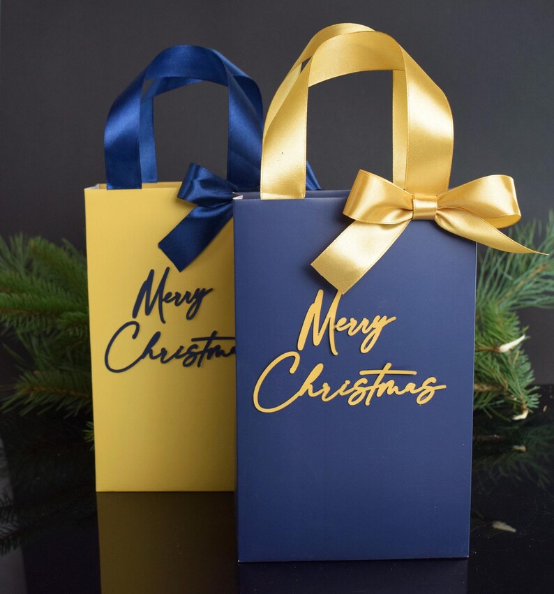 May include: Two gift bags with gold handles and bows. One bag is yellow with a navy blue "Merry Christmas" inscription. The other bag is navy blue with a gold "Merry Christmas" inscription.