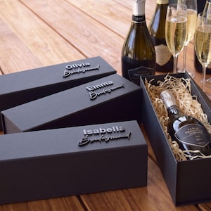 Pode incluir: Três caixas de presente retangulares em preto fosco com fecho magnético, cada uma com letras em relevo que dizem Olivia, Emma ou Isabella seguidas de Bridesmaid. Uma caixa aberta revela uma garrafa de Prosecco sobre palha de madeira.