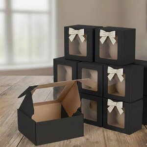 May include: Black gift boxes with clear window panels and cream-colored bows. One box is open, revealing a tan interior. The boxes are stacked on a wooden surface, suggesting a presentation or packaging theme.