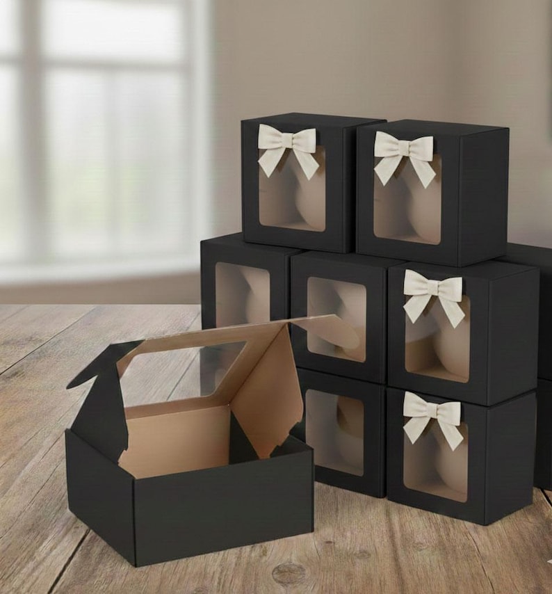 May include: Black gift boxes with clear window panels and cream-colored bows. One box is open, revealing a tan interior. The boxes are stacked on a wooden surface, suggesting a presentation or packaging theme.