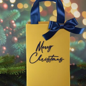 May include: A yellow gift bag with a blue ribbon and the words "Merry Christmas" printed on the front.
