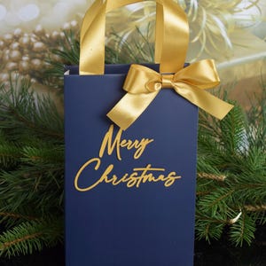 May include: A navy blue gift bag with a gold ribbon and bow. The bag has the words "Merry Christmas" written in gold script.