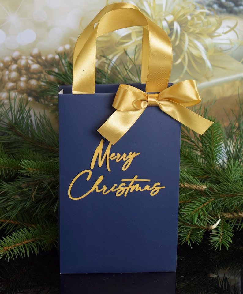 May include: A navy blue gift bag with a gold ribbon and bow. The bag has the words "Merry Christmas" written in gold script.