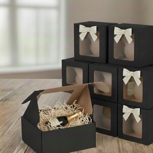 May include: Black gift boxes with clear windows and cream-colored bows are stacked on a wooden surface. One box is open, revealing a bottle of champagne nestled in wood shavings. The boxes are designed for gifting.