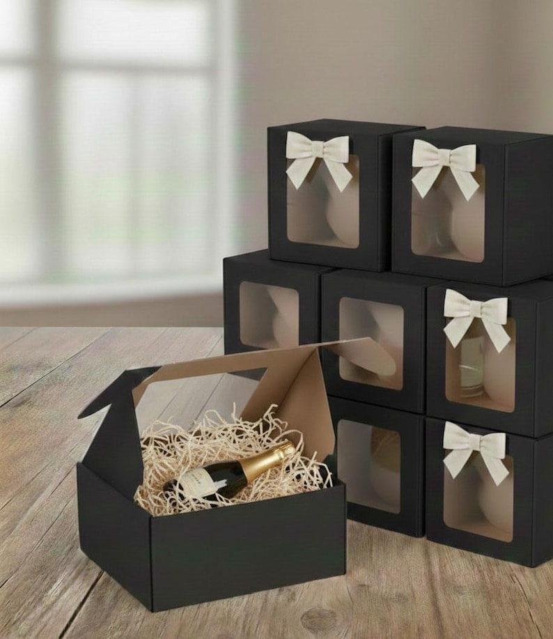 May include: Black gift boxes with clear windows and cream-colored bows are stacked on a wooden surface. One box is open, revealing a bottle of champagne nestled in wood shavings. The boxes are designed for gifting.