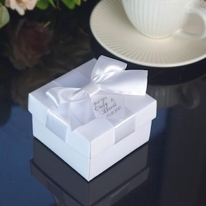 Può includere: Una scatola regalo bianca con un fiocco di raso e un'etichetta con la scritta "Thank You Emily & David 05.12.2020". La scatola è su una superficie riflettente, con una tazza e un piattino bianchi sullo sfondo.