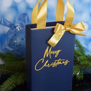 May include: A blue gift bag with a gold ribbon and bow. The bag has the words "Merry Christmas" written in gold on the front.
