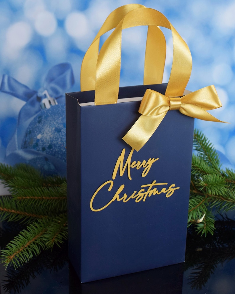 May include: A blue gift bag with a gold ribbon and bow. The bag has the words "Merry Christmas" written in gold on the front.
