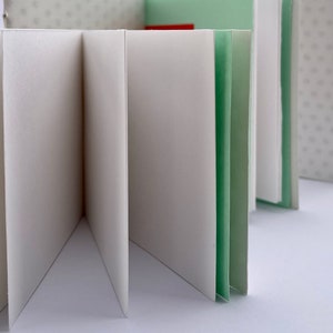 Può includere: Un piccolo quaderno bianco con una copertina verde è aperto per rivelare pagine bianche. Il quaderno è piegato a metà e le pagine sono a ventaglio.