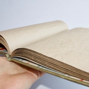 Puede incluir: Un libro abierto con páginas en blanco, la cubierta es una tela estampada con un diseño verde, marrón y blanco. El libro está siendo sostenido abierto por una mano.