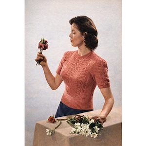 May include: A vintage-style, short-sleeved, coral-colored knit top with a textured leaf pattern. The top is paired with a dark blue skirt. The image shows a woman holding a small bouquet of flowers.