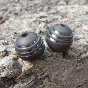 May include: Two small, spherical, metal objects with a dark gray finish. Each sphere has a series of horizontal grooves and a small hole at the top. The objects are resting on a rough, gray stone surface.