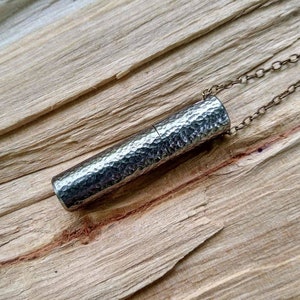May include: A silver necklace with a hammered metal cylinder pendant. The pendant is attached to a silver chain.