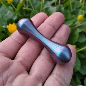 Könnte beinhalten: Ein lila-grüner Metall-Fidget-Spinner mit einer glatten, abgerundeten Form. Der Spinner wird in einer Hand gehalten.