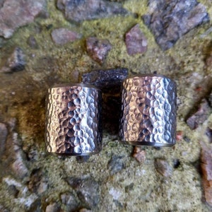 May include: Two silver-colored, hammered metal cylinders. The cylinders have a textured surface with a pattern of small, irregular indentations. The cylinders are set against a background of rough, multi-colored stones.