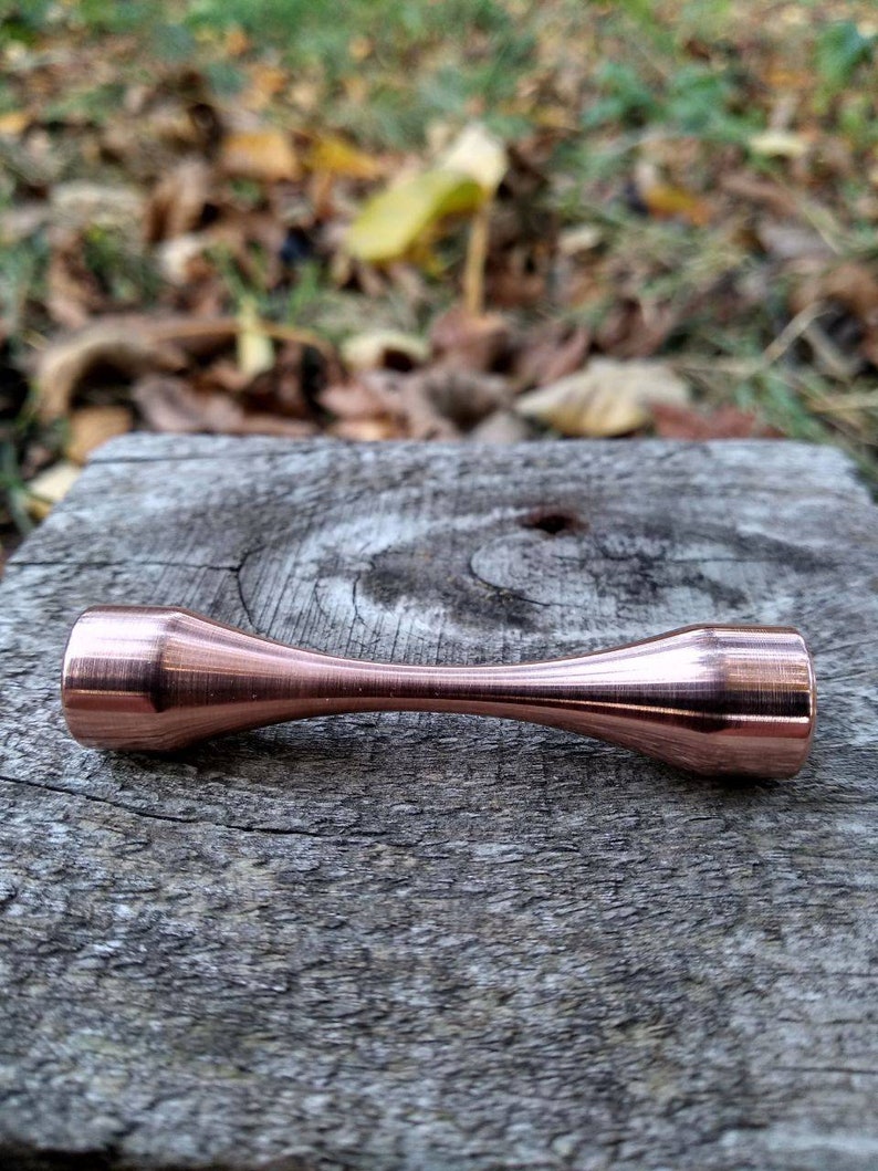 Puede incluir: Una varilla de metal de color cobre con extremos redondeados, que se encuentra sobre una pieza de madera desgastada.