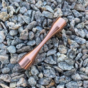 Puede incluir: Una peonza de metal de color cobre con una forma redondeada de reloj de arena. La peonza est&aacute; apoyada sobre un lecho de peque&ntilde;as piedras grises y blancas.