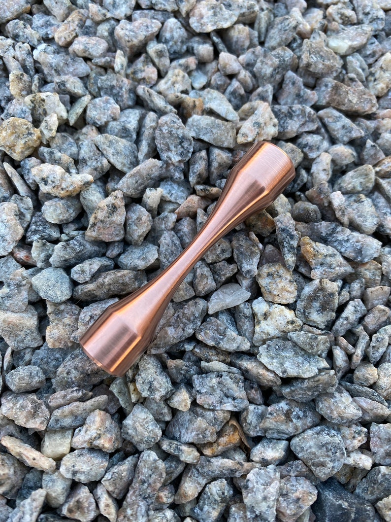 Puede incluir: Una peonza de metal de color cobre con una forma redondeada de reloj de arena. La peonza est&aacute; apoyada sobre un lecho de peque&ntilde;as piedras grises y blancas.