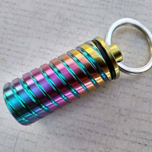 May include: A multi-colored metal keychain with a cylindrical container and a silver ring. The container has a rainbow iridescent finish with alternating bands of green, blue, purple, and gold.
