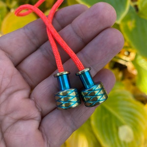 May include: Two small, decorative metal beads with bright orange cords. The beads have a gold-colored base with blue accents and a crisscross design. The cords are a vibrant orange color. The background is blurred with green and yellow foliage.