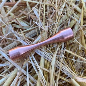 Puede incluir: Una varilla de metal de color cobre con una forma redondeada de reloj de arena. La varilla est&aacute; sobre un lecho de paja.