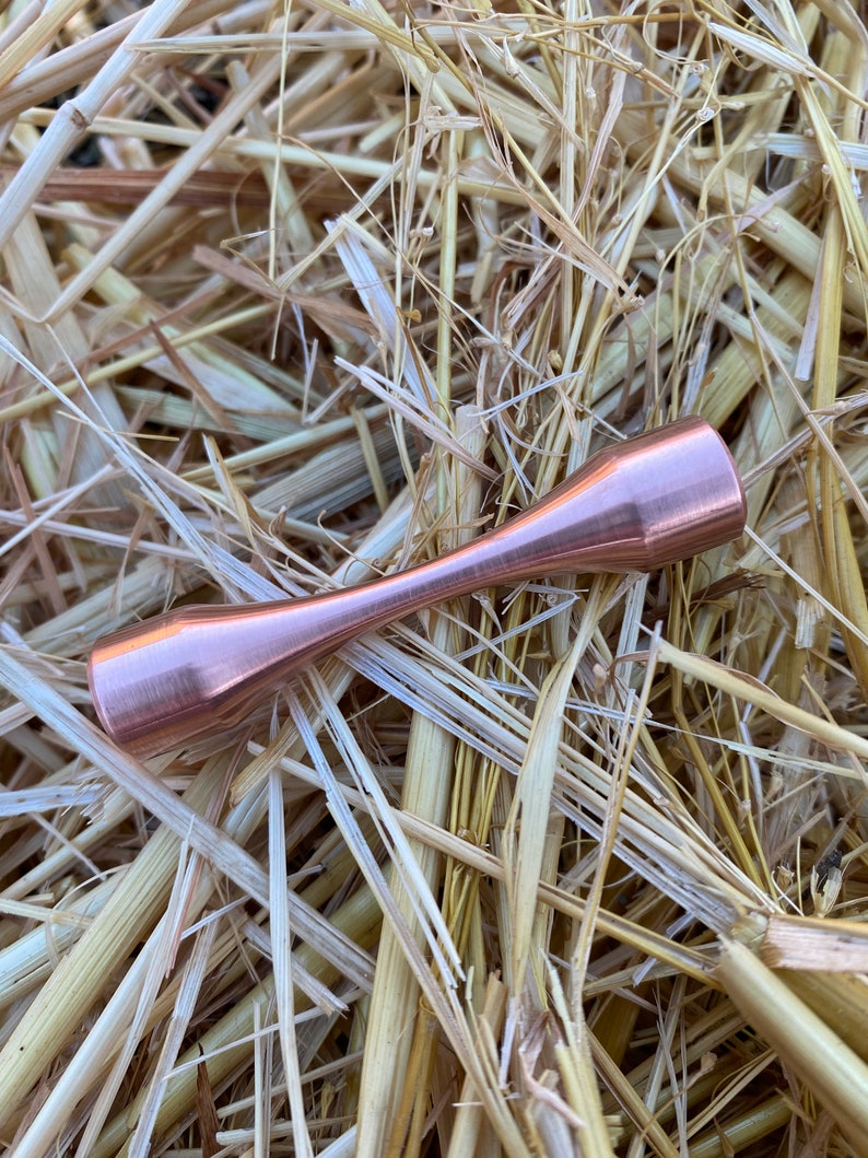 Puede incluir: Una varilla de metal de color cobre con una forma redondeada de reloj de arena. La varilla est&aacute; sobre un lecho de paja.