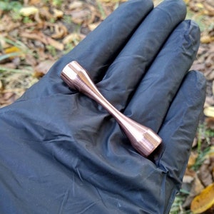 Puede incluir: Primer plano de una varilla de metal de color cobre con extremos redondeados. La varilla se sostiene en una mano enguantada.