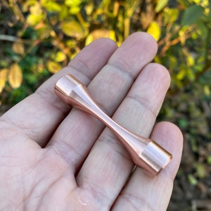 Puede incluir: Un juguete fidget de metal de color oro rosa con forma de mancuerna. El juguete mide aproximadamente 5 cm de largo y tiene un acabado liso y pulido.