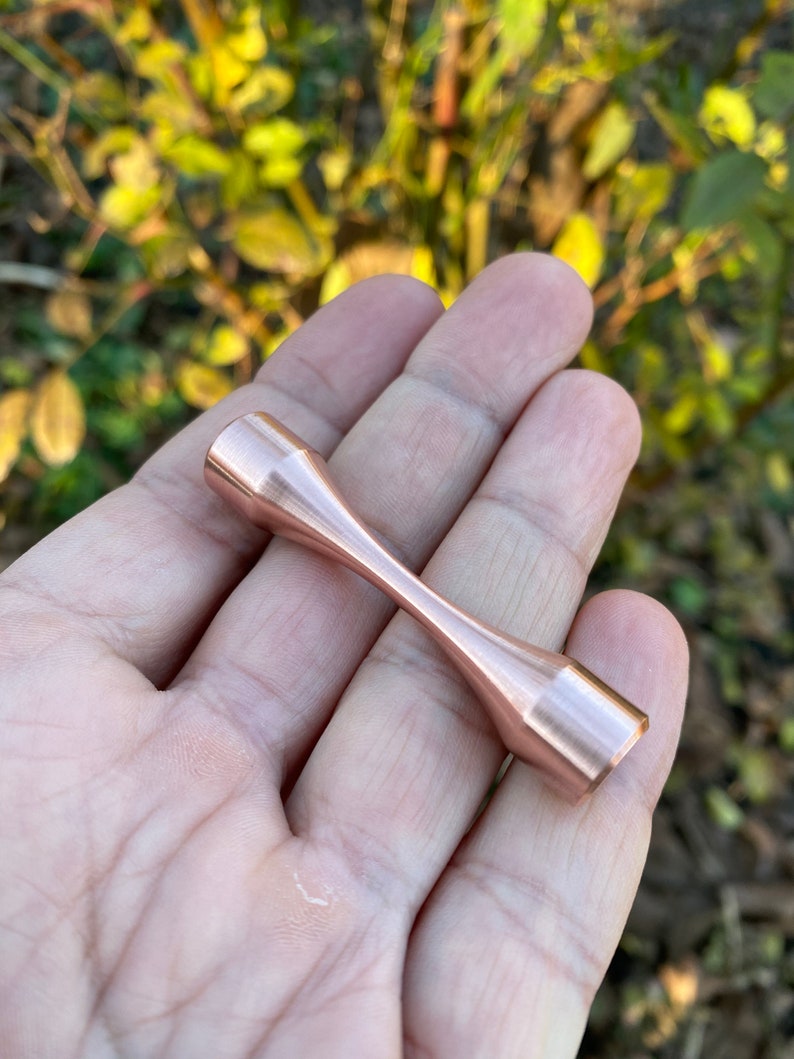 Puede incluir: Un juguete fidget de metal de color oro rosa con forma de mancuerna. El juguete mide aproximadamente 5 cm de largo y tiene un acabado liso y pulido.