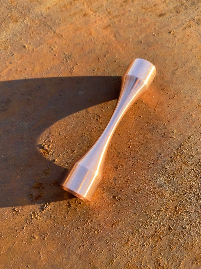 Puede incluir: Un esp&eacute;cimen de prueba de tracci&oacute;n de metal de color cobre con un acabado liso y pulido. El esp&eacute;cimen tiene la forma de una mancuerna con una secci&oacute;n central estrecha y extremos m&aacute;s anchos.