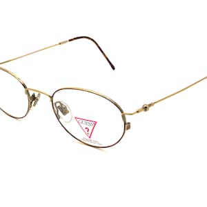 Könnte beinhalten: Goldfarbene Metallbrille mit ovalen Gläsern und einem Guess-Logo auf der rechten Linse.