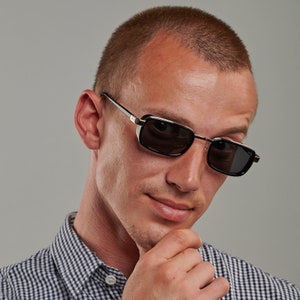 May include: A man wearing a black and silver framed sunglasses with black lenses. He is wearing a blue and white checkered shirt and has a short haircut.