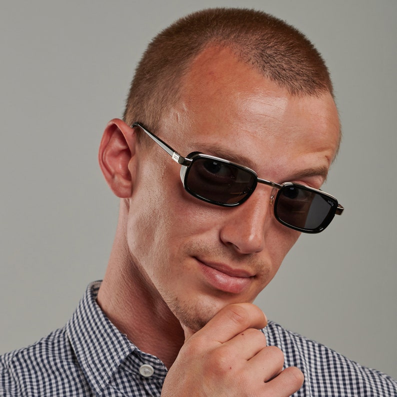 May include: A man wearing a black and silver framed sunglasses with black lenses. He is wearing a blue and white checkered shirt and has a short haircut.