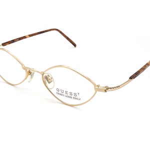 May include: Gold-tone Guess eyeglasses with a cat-eye shape. The frames have a thin, delicate design with a subtle oval lens shape. The temples feature a tortoiseshell pattern. The text "GUESS? DEMO LENS ONLY" is printed on the lens.