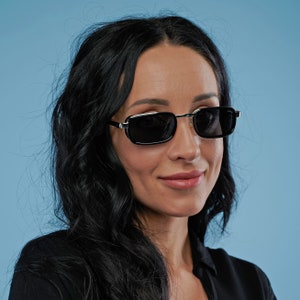 May include: A woman with long black hair wearing black sunglasses with a silver frame. She is wearing a black shirt and has a neutral expression.