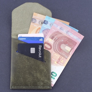 Op de afbeelding: Olijfgroene leren portemonnee met flap, met eurobiljetten en creditcards. De portemonnee heeft een minimalistisch ontwerp met zichtbare stiksels. De kaarten bevatten een "business" kaart en een "Revolut" kaart.