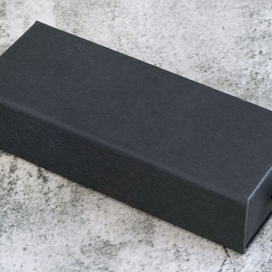 May include: A black rectangular box with a textured surface and a black ribbon pull tab.