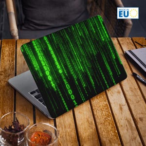 May include: Laptop skin with a green binary code design on a black background. The laptop is on a wooden table, with a notebook and pen. The skin is labelled "MADE IN EU".