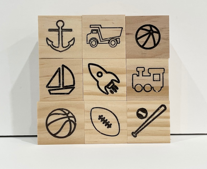 May include: Nine wooden blocks with black line drawings of various objects, including an anchor, a truck, a basketball, a sailboat, a rocket, a train, a football, a baseball and bat, and a basketball.