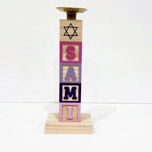 Könnte beinhalten: Ein Kerzenhalter aus Holz mit einem Davidstern auf dem oberen Block und dem Namen "SAMI" auf den Blöcken darunter. Die Blöcke sind in Rosa, Lila und Blau bemalt.