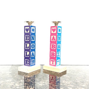 May include: Two wooden candle holders shaped like stacked blocks. The blocks spell out "ELOHAH" and "AMABXY" in blue and pink letters. Each holder has a silver candle cup on top.