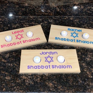 May include: Three wooden candle holders with a Star of David and the words "Shabbat Shalom" in different colors. The names "Leat", "Rachel", and "Jordyn" are written on each holder.