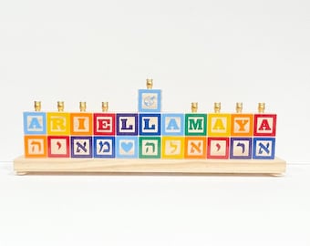 Può includere: Una menorah di legno con un arcobaleno di colori e il nome "ARIEL LAMAYA" scritto in blocchi. La menorah ha un blocco centrale con una trottola su di esso.