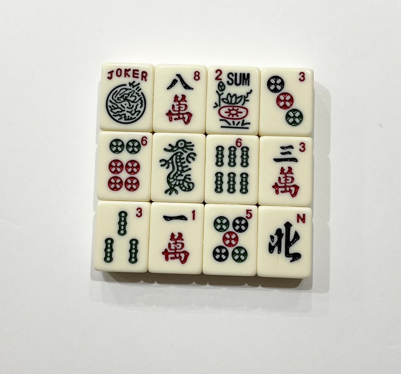 Op de afbeelding: Een set van 12 witte mahjong-tegels met zwarte en rode Chinese karakters en symbolen. De tegels zijn gerangschikt in een 3x4 raster. De tegels bevatten de nummers 1, 2, 3, 5, 6, 8 en de karakters "Joker", "Som", "Noord" en "Draak".