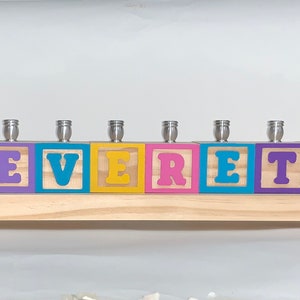 May include: A wooden menorah with a Star of David on the top block and the name EVERETT spelled out on the other blocks. Each block has a different color and a small animal or design on it. The menorah is on a wooden base.