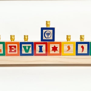 May include: A wooden menorah with a personalised name "LEVI" spelled out in colourful blocks. Each block features a different Hebrew symbol or letter. The menorah has nine candle holders, with a central shamash holder.