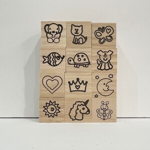 May include: A set of 12 wooden stamps with various cute animal and object designs. The stamps are arranged in a grid pattern and feature images such as a fish, a turtle, a unicorn, a butterfly, a heart, a crown, a moon, a sun, and a dog.