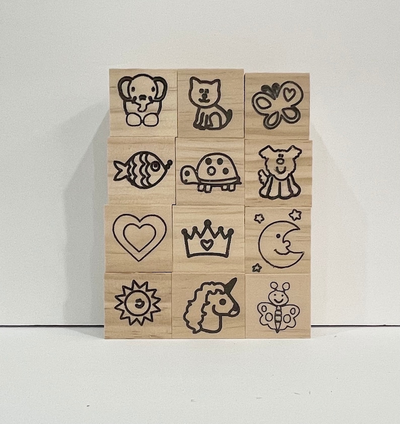 May include: A set of 12 wooden stamps with various cute animal and object designs. The stamps are arranged in a grid pattern and feature images such as a fish, a turtle, a unicorn, a butterfly, a heart, a crown, a moon, a sun, and a dog.
