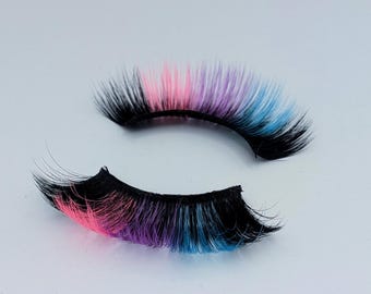 May include: A pair of colorful false eyelashes. The lashes are black at the base, transitioning to pink, purple, and blue at the tips. The lashes are arranged on a white surface.